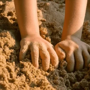 Two child's hands are playfully digging into soft, golden sand. The sunlight casts warm tones, evoking a feeling of joy and relaxation.